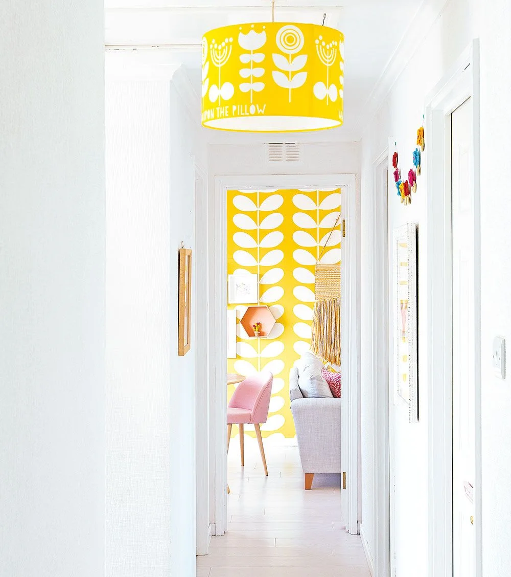 A white hallway with a yellow floral pendant looks toward a room with bold yellow leaf wallpaper, a gray sofa, pink chair and pale floor.