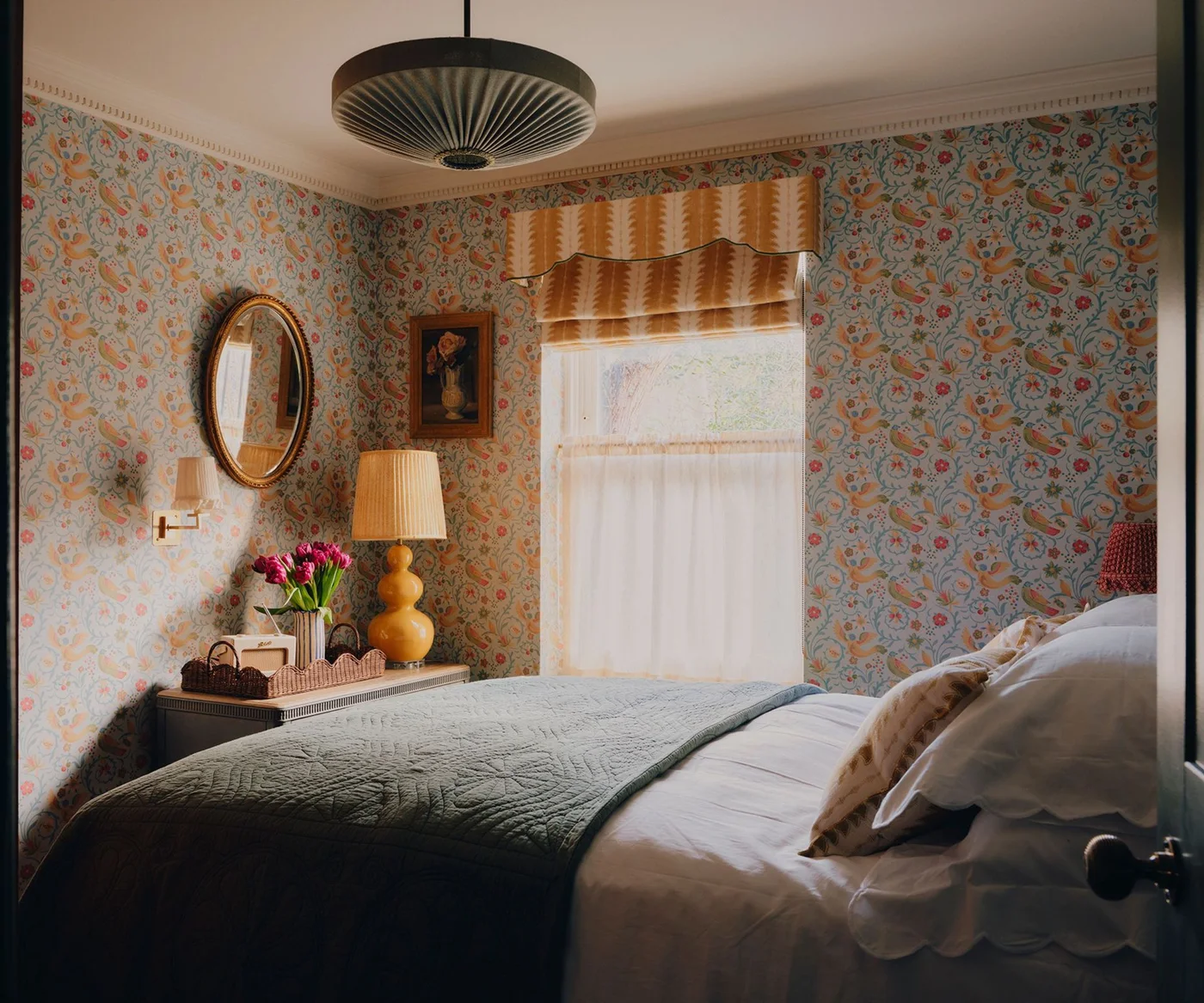 Dense blue floral wallpaper surrounds pale blue-gray bedding beside a yellow-and-orange patterned window treatment and a warm-colored lamp