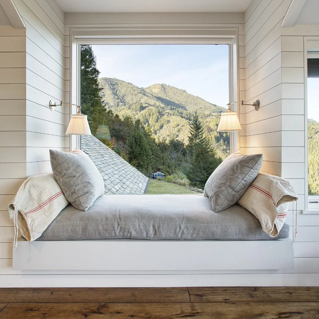A pale cushioned window seat between white board walls with swing arm lamps and a mountain view