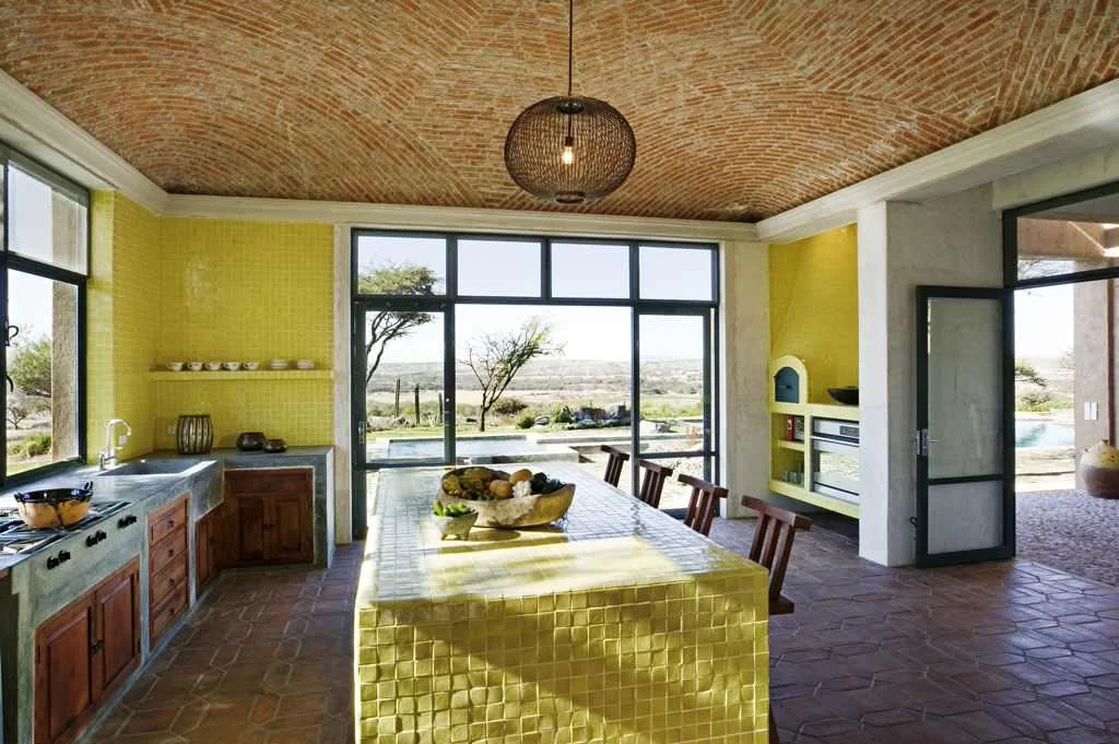 Yellow-green tiled island and wall in a kitchen with wood cabinets, dark window frames and a brick ceiling.