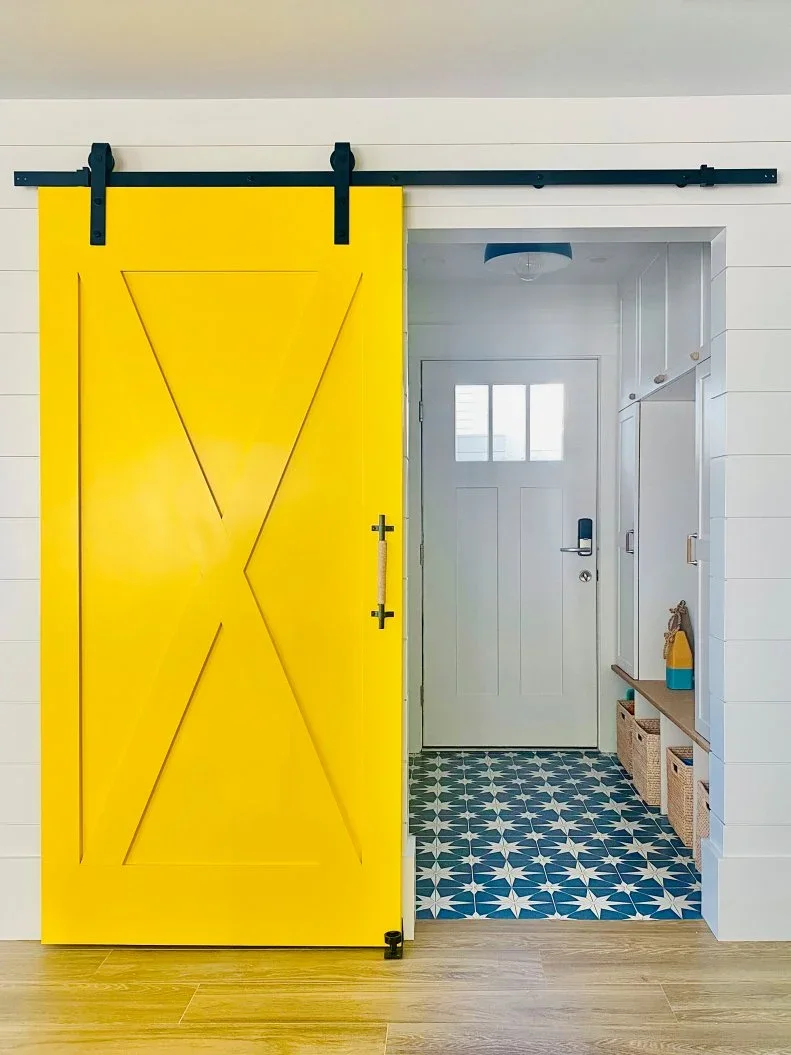 A wood-topped bench with woven basket bays sits inside a white mudroom behind a bright yellow sliding door.