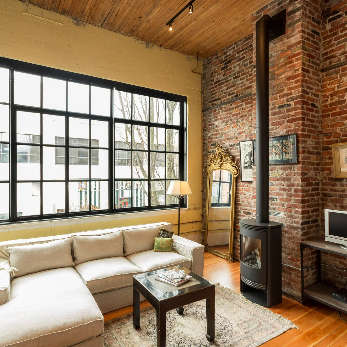 A narrow black stove and tall pipe stand against a red brick loft wall beside large gridded windows, a pale sofa and an ornate mirror.