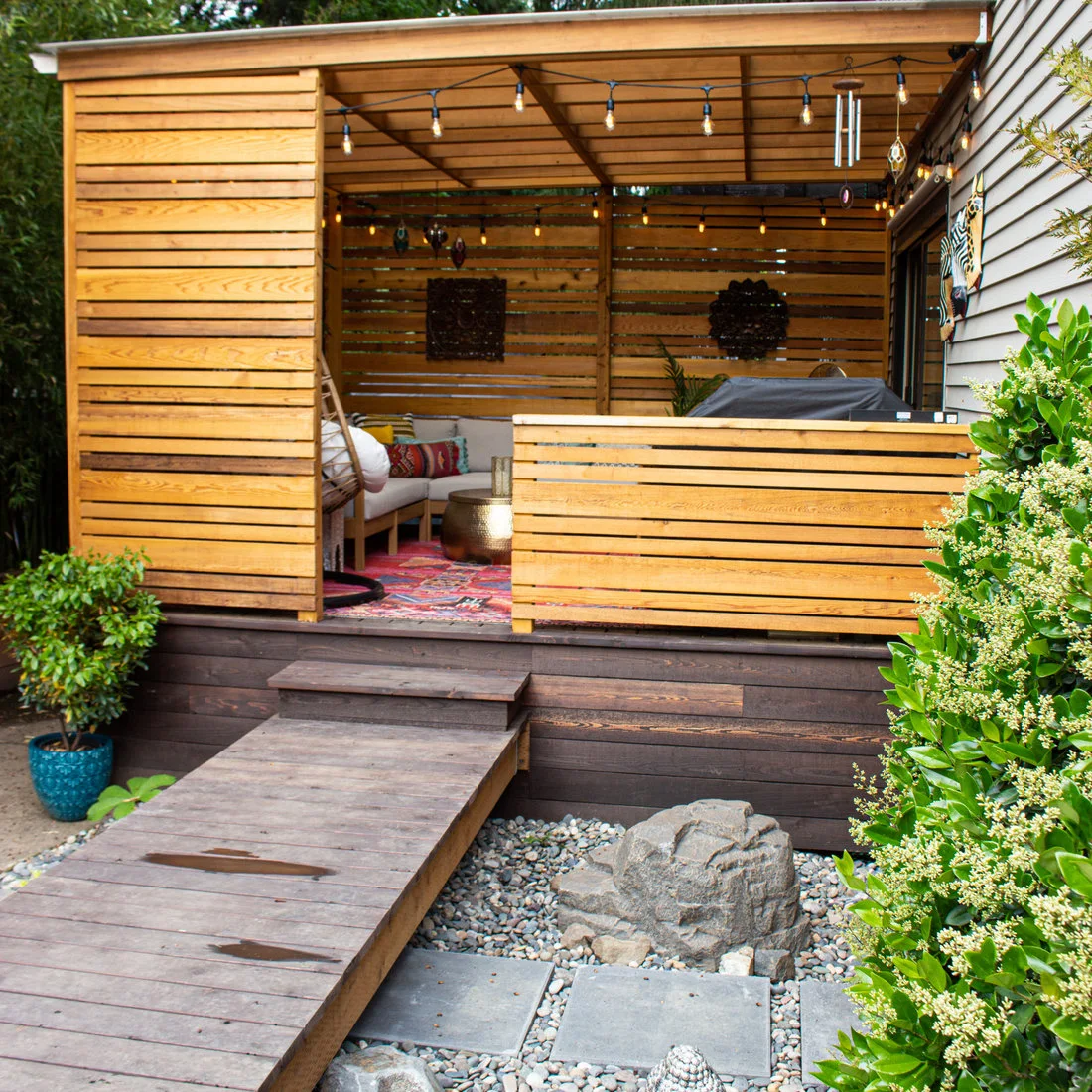 A timber-roofed garden shelter has slatted side walls, a sofa and an egg chair on a stand