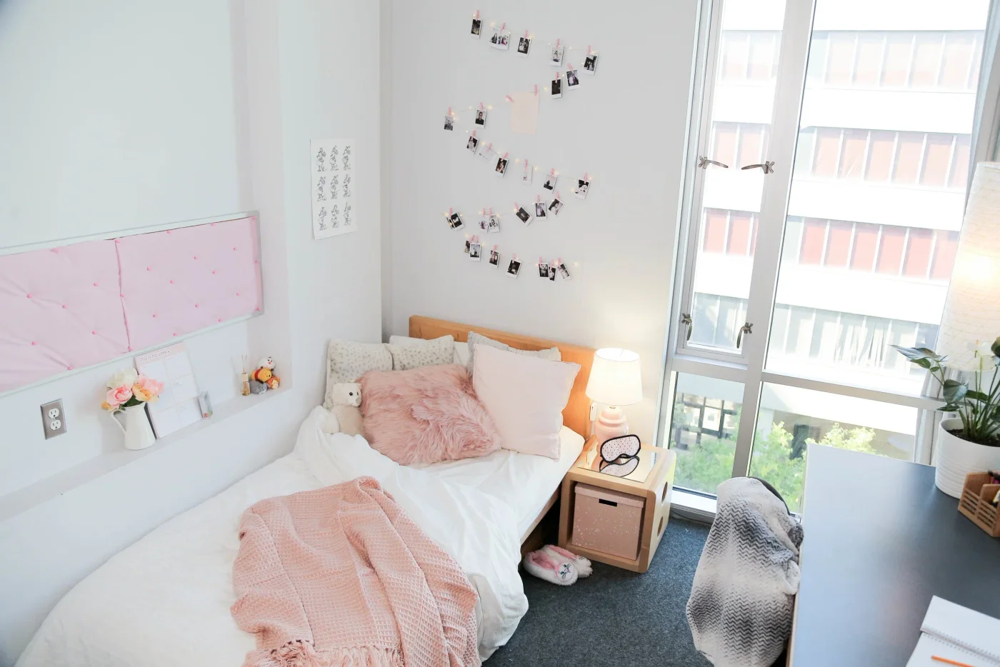 Narrow student bed with white bedding, pink textured throw, small bedside cube and photos on the wall