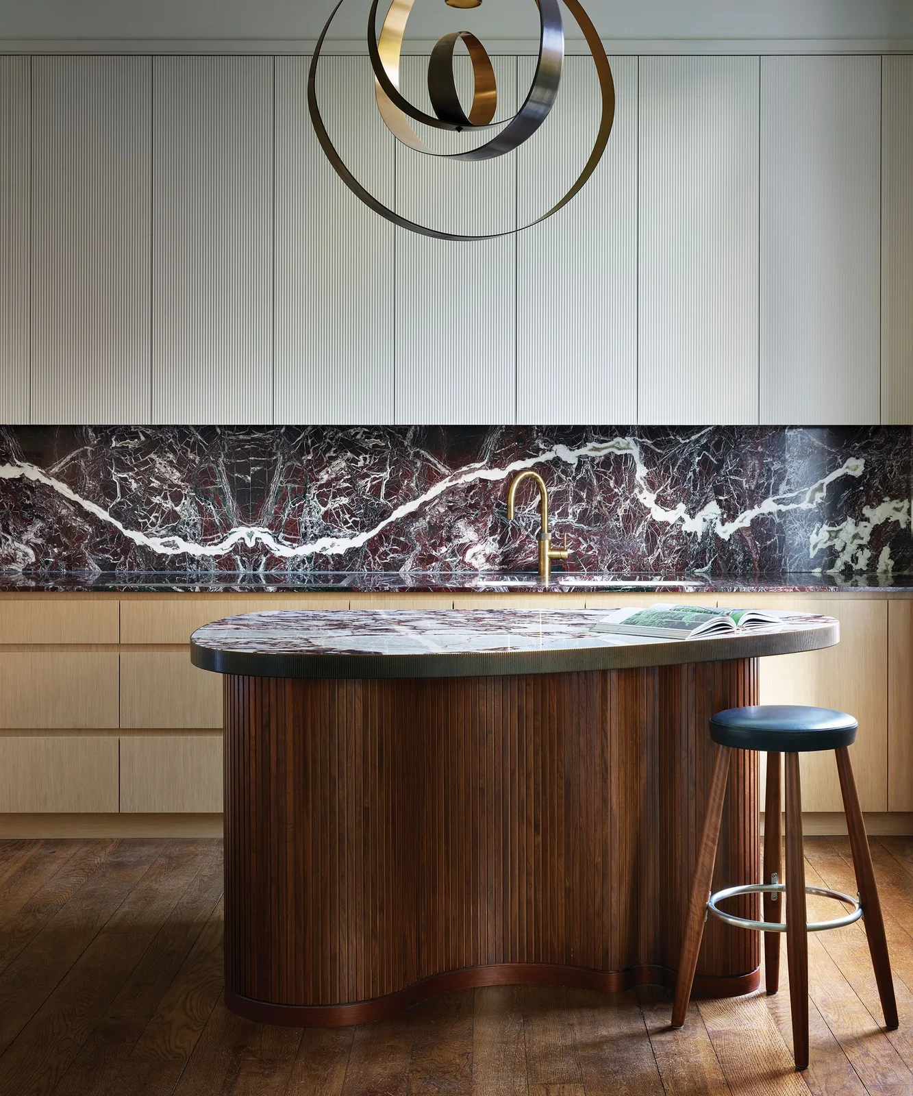 A curved fluted walnut island stands before pale cabinets, a deeply veined dark backsplash and a gold-colored faucet.
