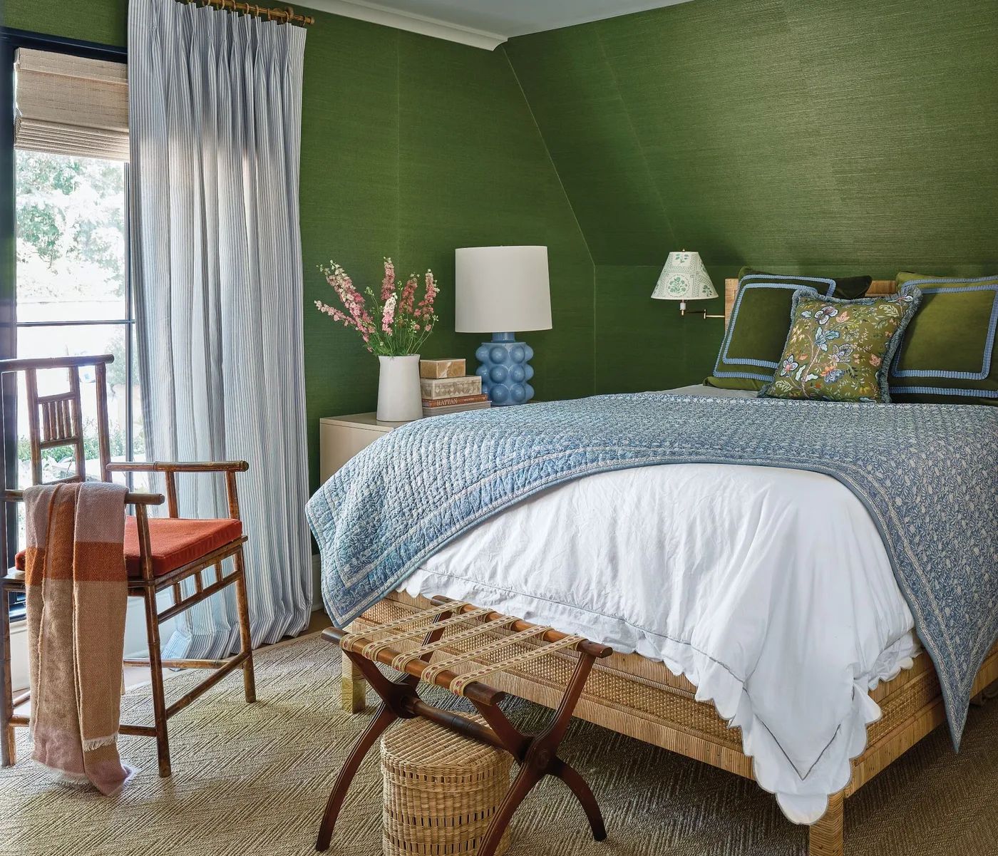 Green textured wallpaper around a bed with a blue quilt, pale curtains and a wooden accent chair.