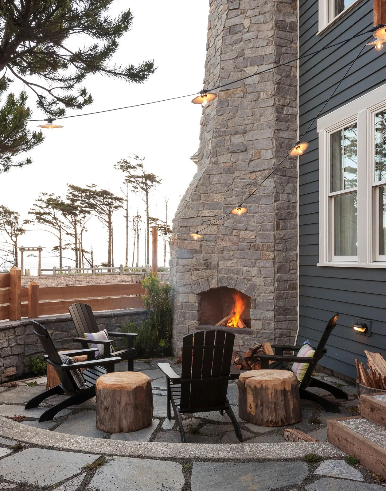 A tall stone outdoor fireplace faces dark timber chairs and stump tables on a paved patio.