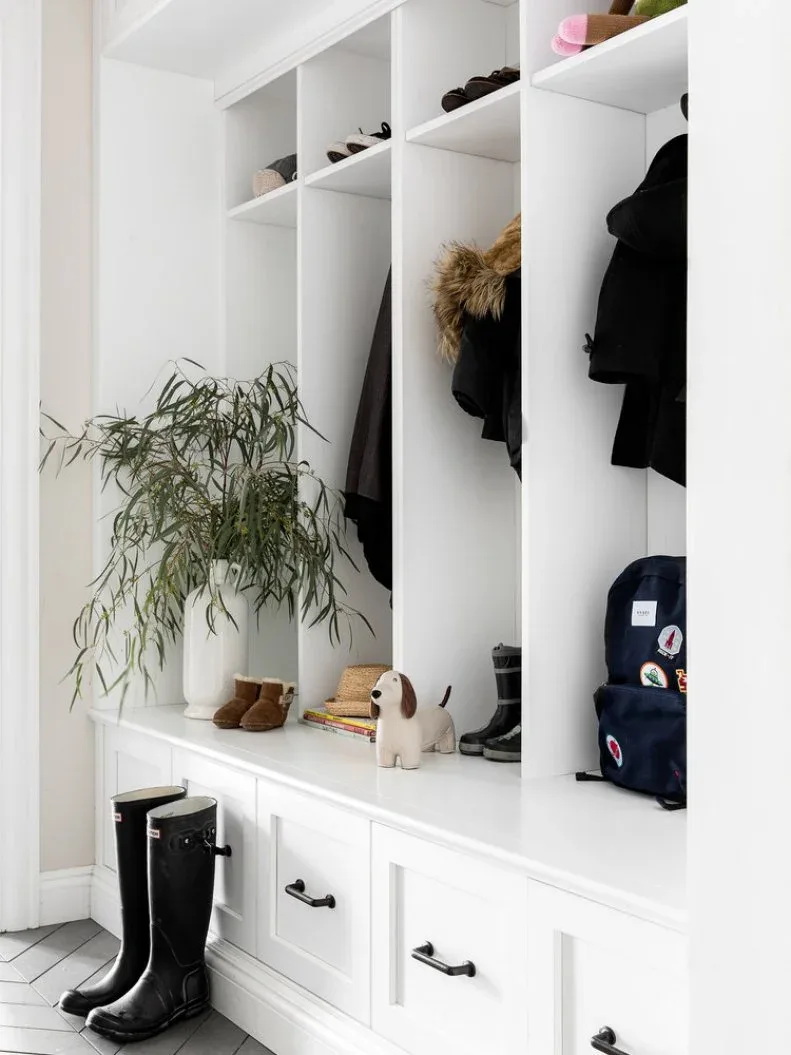 White divided entry bench with black handles, lower drawers, shoe and coat bays, bags and boots on pale floor