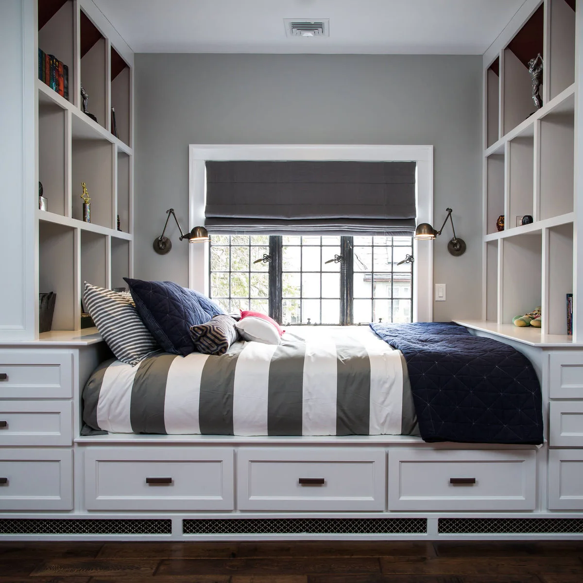 Built-in boy’s bed beneath a window with pale bookshelves, lower drawers and blue-gray bedding
