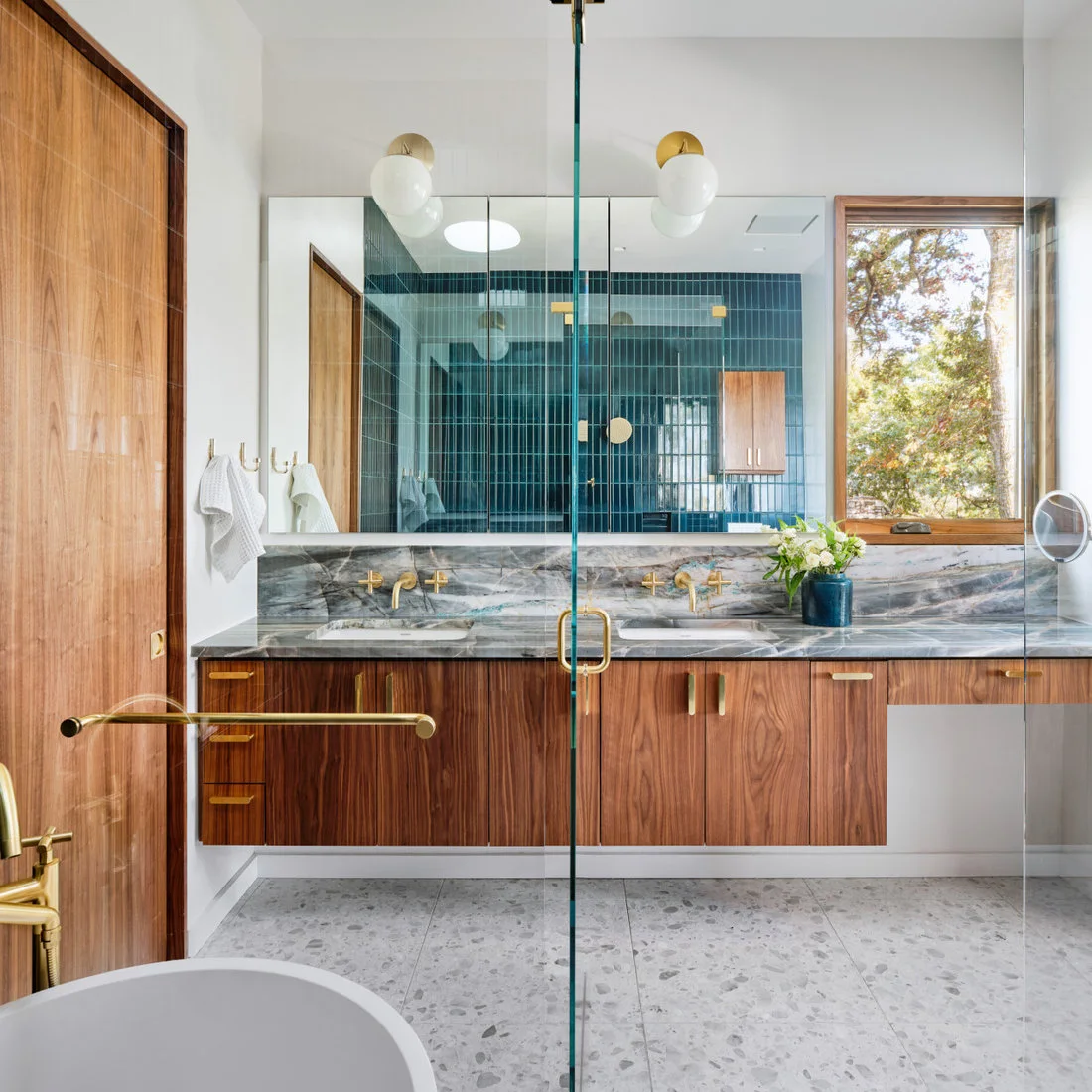 Pale terrazzo lies below a timber vanity with a veined counter and a mirror reflecting blue shower tiles.
