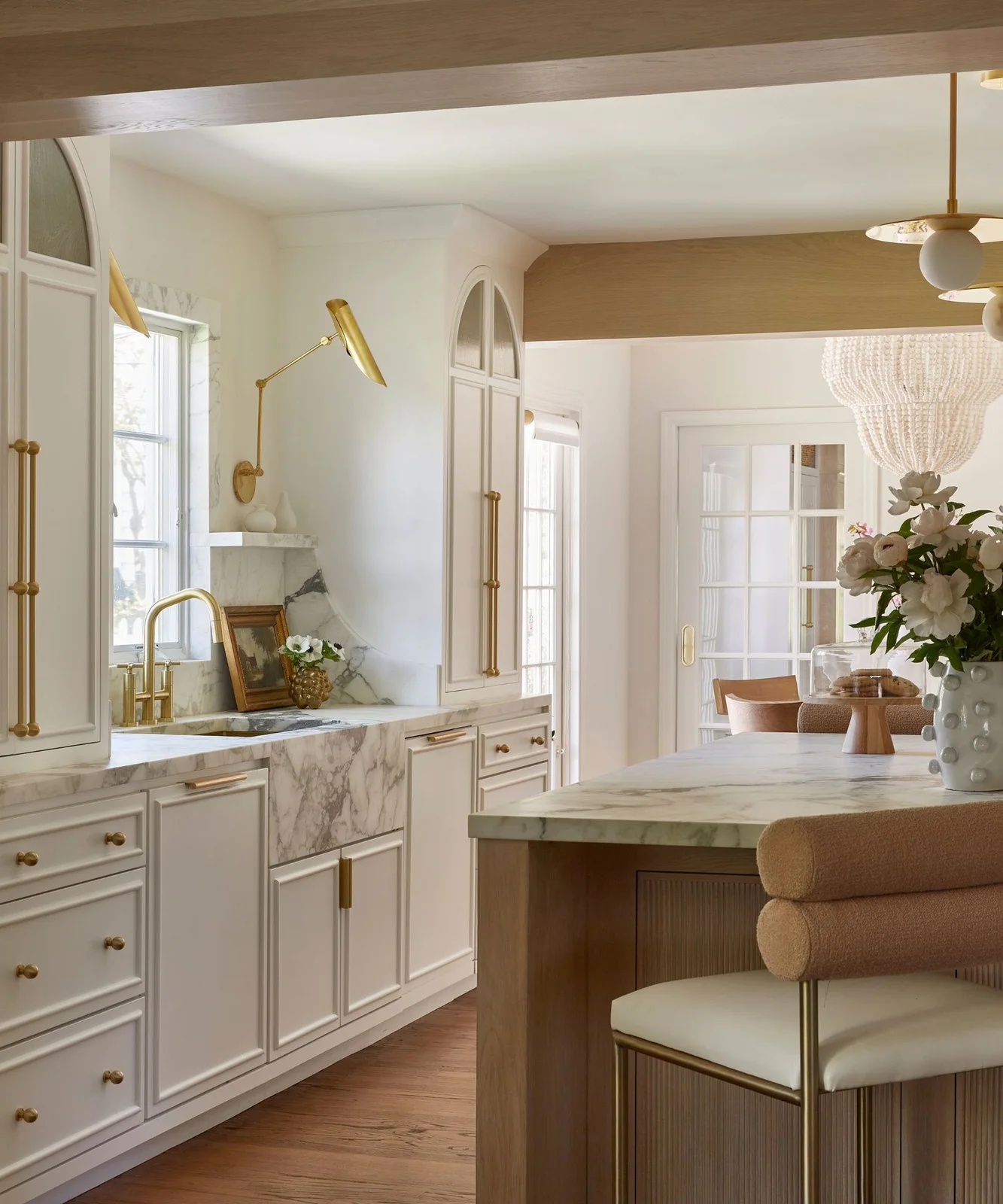 White upper cabinets with arched detailing flank a window above a broad veined sink and brass-colored hardware.