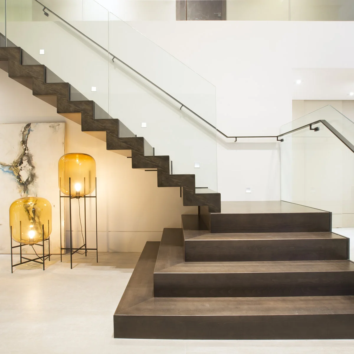 Modern dark staircase with glass guards, small wall lights and a continuous dark handrail around the landing