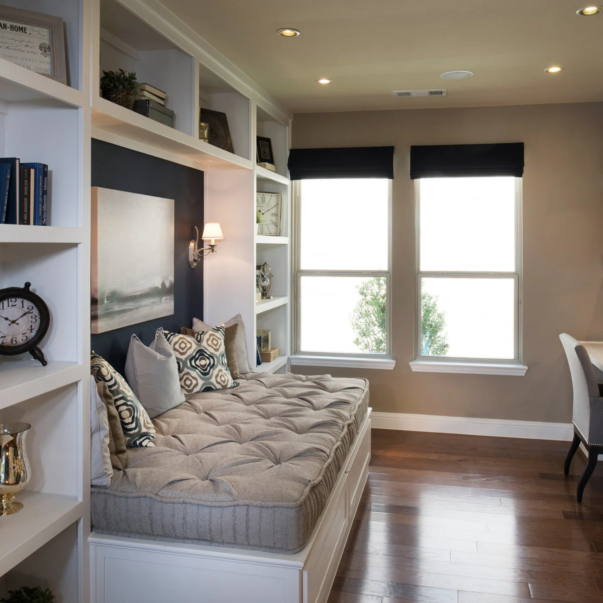 A broad tufted cushion sits on a white daybed base between shelves, with a dark wall recess and two tall windows nearby.