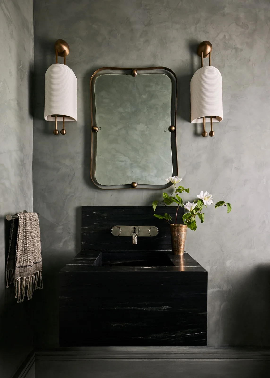 A dark wall-mounted rectangular basin sits below a rounded rectangular mirror between two pale cylindrical wall lights against a textured gray wall