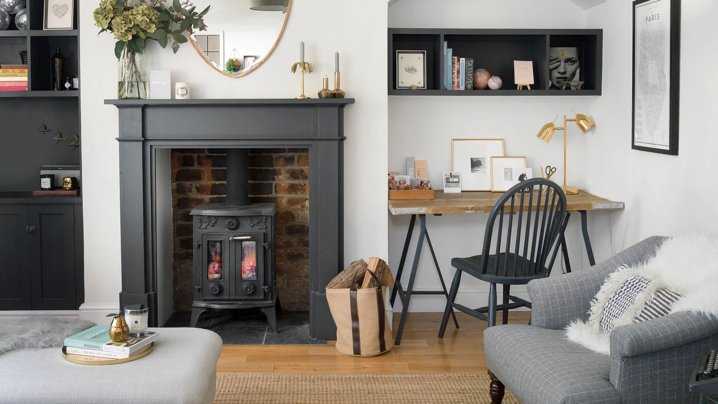A dark mantel and stove sit against white walls beside a compact wood desk, black chair and shelves, with gray seating and a woven rug in the foreground.