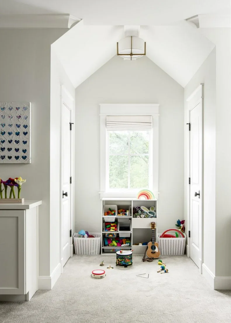 A white vaulted alcove holds low cube toy storage beneath a window, two white baskets and small instruments, with doorways on both sides.