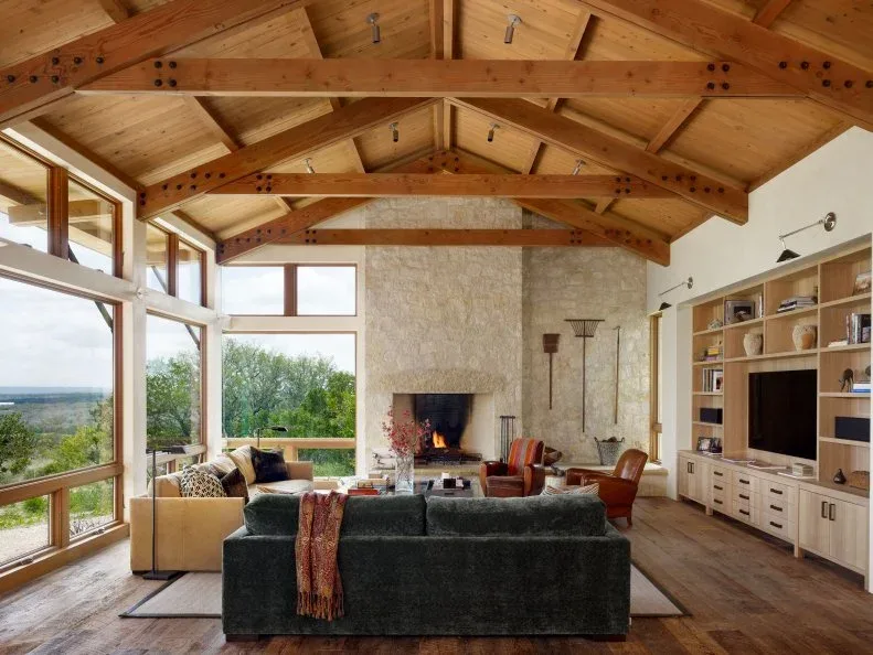 A tall pale stone fireplace rises beneath timber roof beams beside large windows and open living-room shelving.