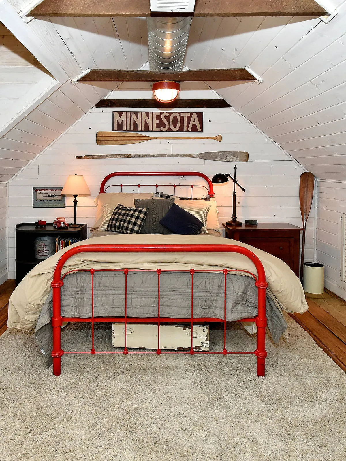 A red metal bed anchors a pale attic bedroom beneath angled ceiling beams.