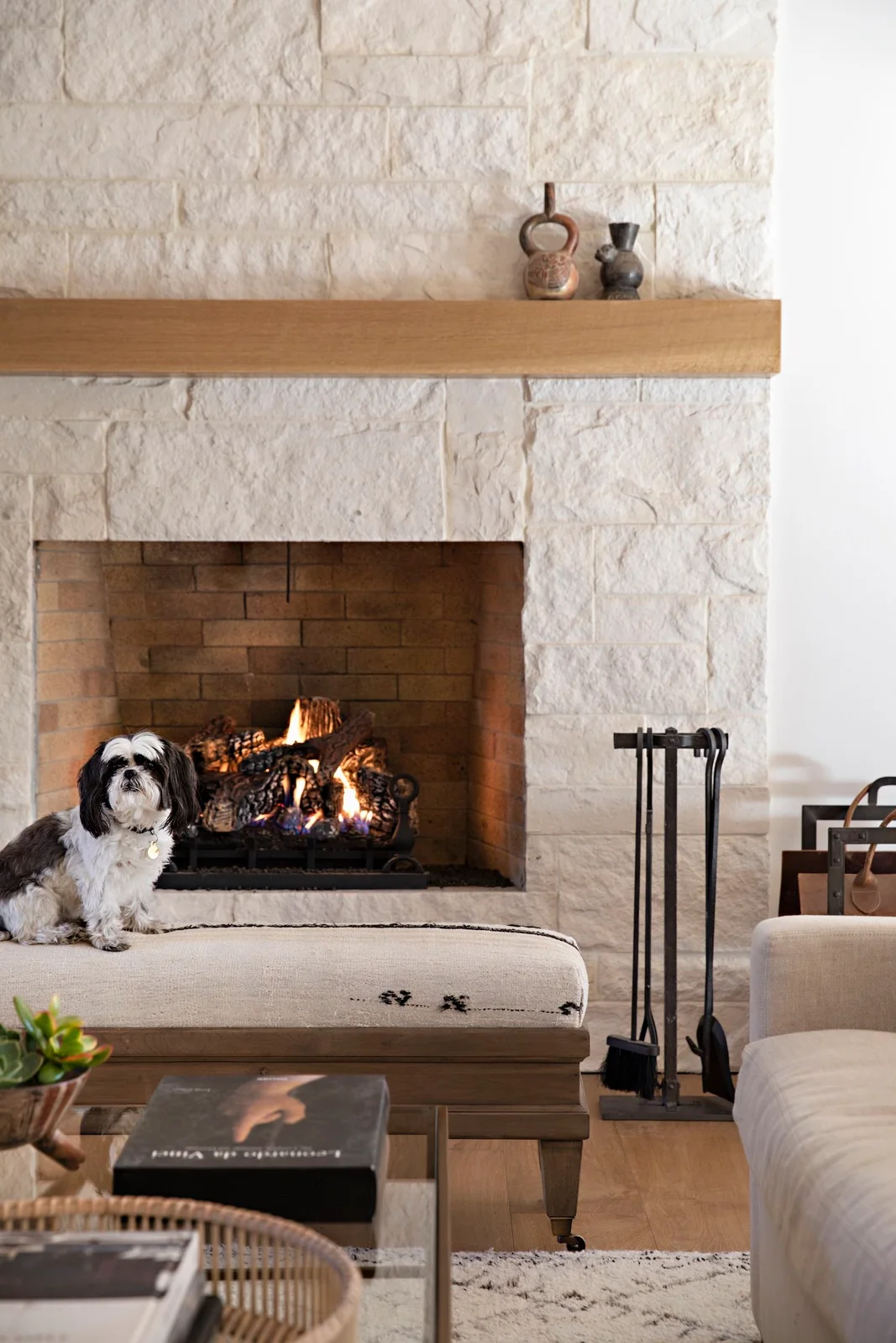 A pale rough-stone fireplace has a plain timber mantel above a brick-lined opening and upholstered bench.