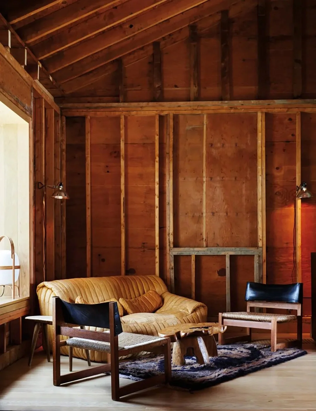 A pale tan leather sofa sits against exposed wooden framing beside two dark-backed chairs and an irregular low table