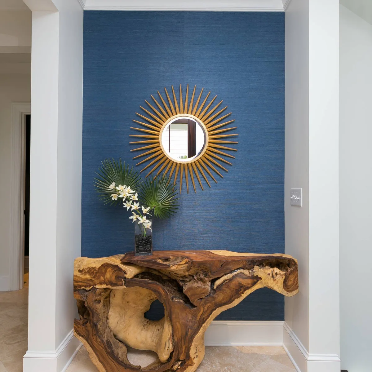A sculptural wood console against a blue wall with a sunburst mirror and one tall flower arrangement.