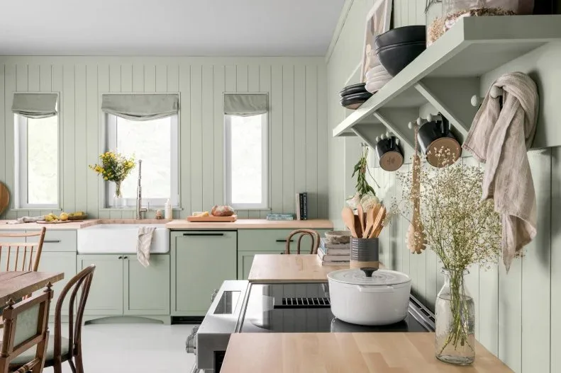 Soft green kitchen with vertical wall boards, wood counters, open shelves, hanging pans and dining chairs beneath tall windows