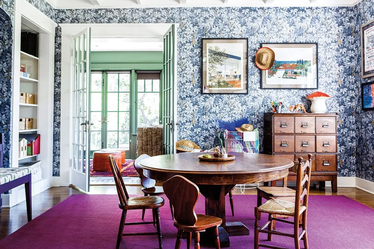 Framed pictures above a small drawer cabinet on blue floral wallpaper beside a round dining table