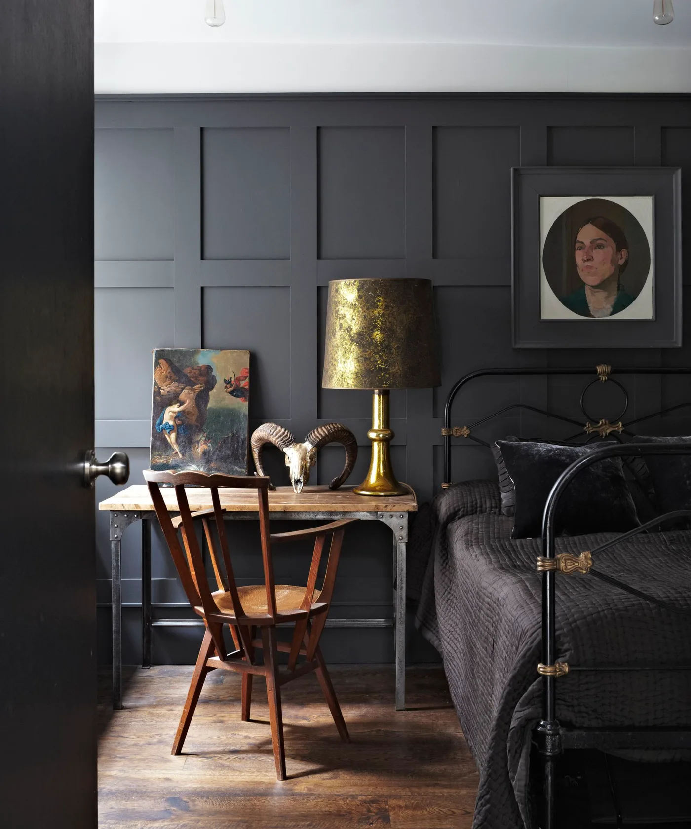 Charcoal paneled bedroom with a dark metal bed, wooden chair, desk-style bedside surface, portrait, and gold-colored lamp.