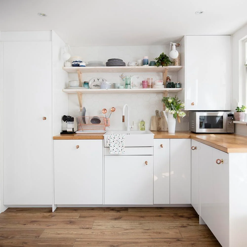A white kitchen has wood counters, two open shelves of dishes and glasses, an apron-front sink, small warm metal knobs and tall closed storage at one side.