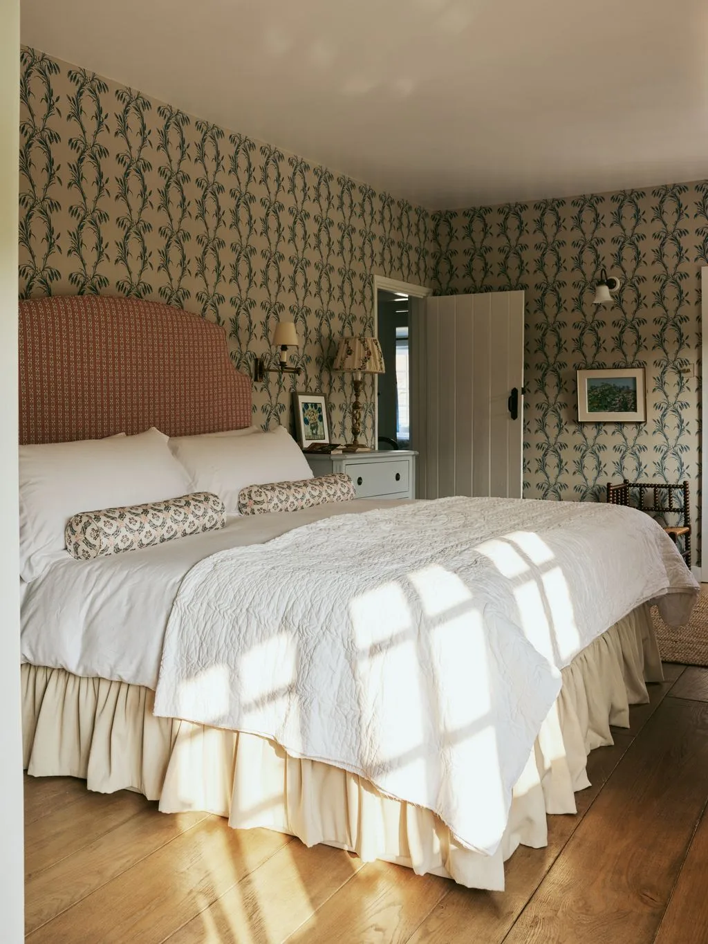 Bedroom with green botanical wallpaper, reddish patterned headboard, white bedding, pale bedside drawers and wood floor