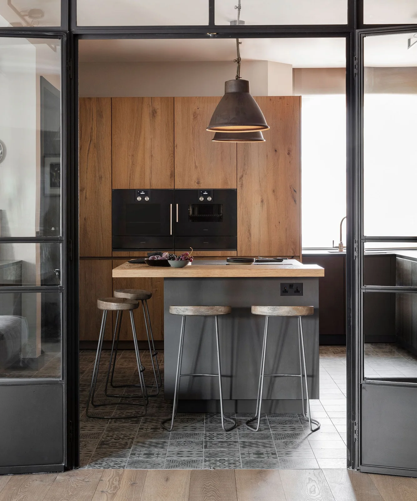 A vintage oak cabinet wall with black ovens stands behind a gray island and simple stools, seen through black framed glass doors.