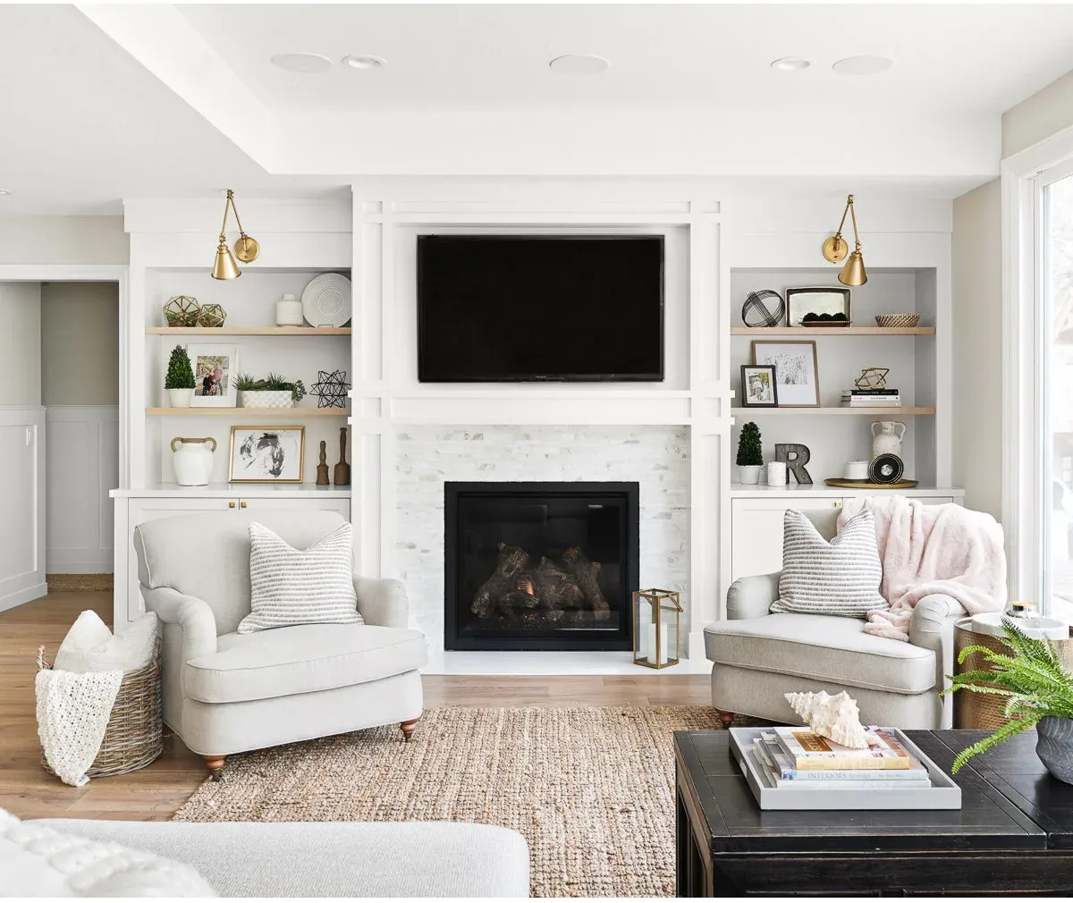 A television above a white fireplace between pale wood shelves, brass-colored sconces and two cream chairs.