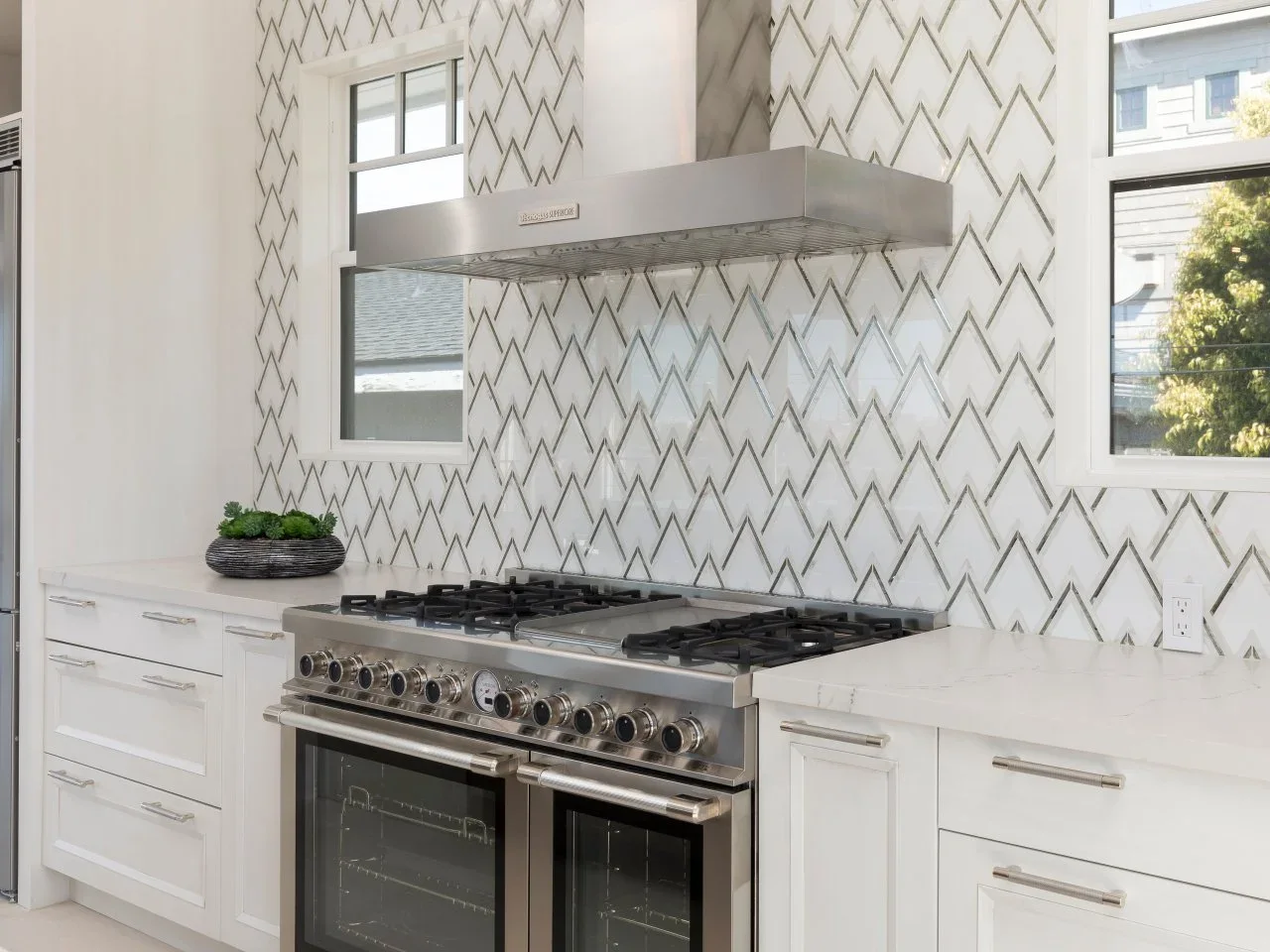 A white kitchen backsplash with outlined geometric shapes rises around two windows behind a range and stainless hood.