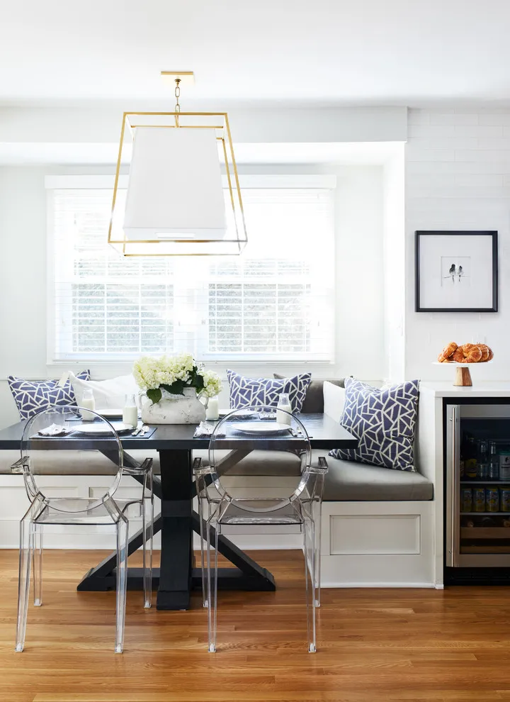 A plain gray bench with patterned cushions sits beneath a window opposite clear dining chairs