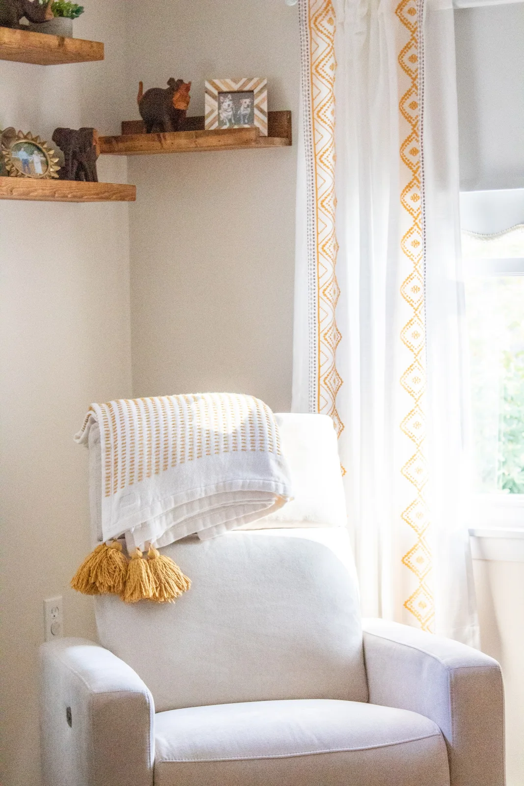 A pale nursery recliner has a mustard-tasseled throw beside patterned curtains and shelves holding animal figures.