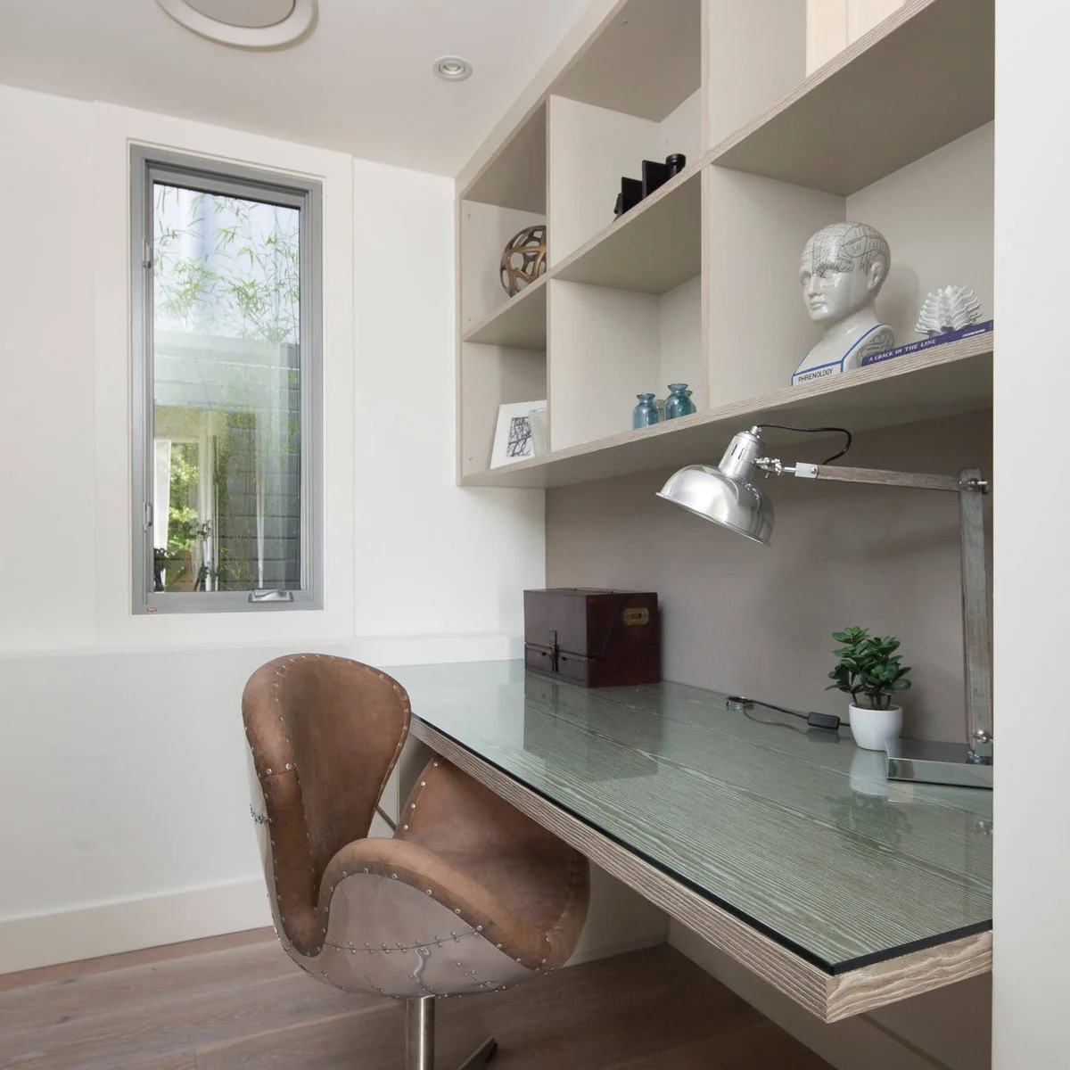 Compact desk with a glass-covered top, brown curved chair, adjustable metal lamp and open wall shelves