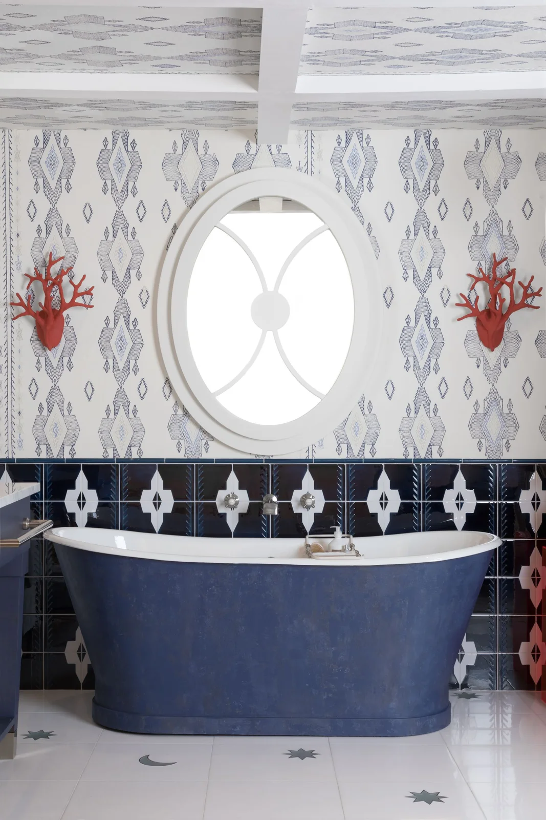 A blue freestanding bath sits beneath a round window between patterned dark lower tiles, finer upper-wall pattern and red wall lights