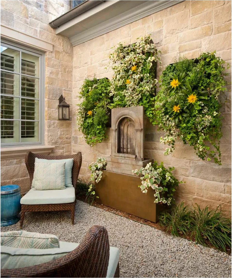 A pale cushioned woven chair sits beside a wall fountain beneath rectangular planted panels on a pale masonry wall.