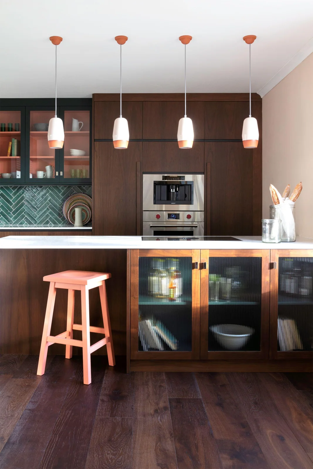 Walnut-framed reeded glass doors line an island beside a coral stool, with walnut tall cabinets and built-in ovens behind.