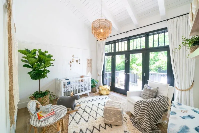 White nursery with crib, adult armchair, geometric pale rug, woven containers, small chairs, textured pendant and black-framed doors.