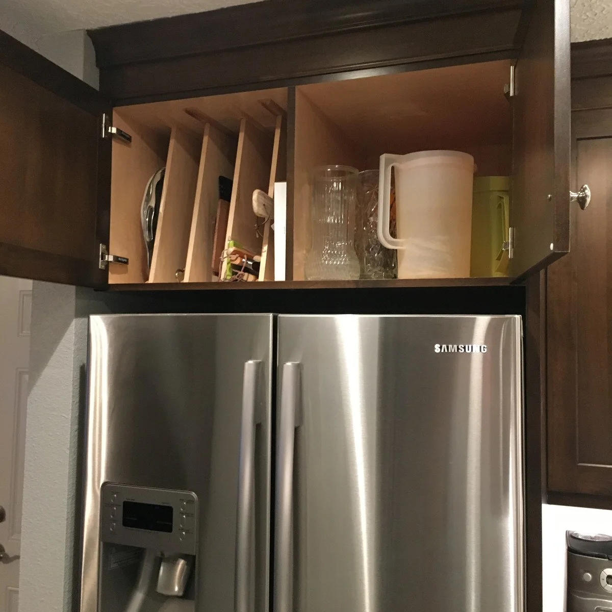 An open dark cabinet above a stainless refrigerator has vertical tray slots on the left and pitchers on the right.