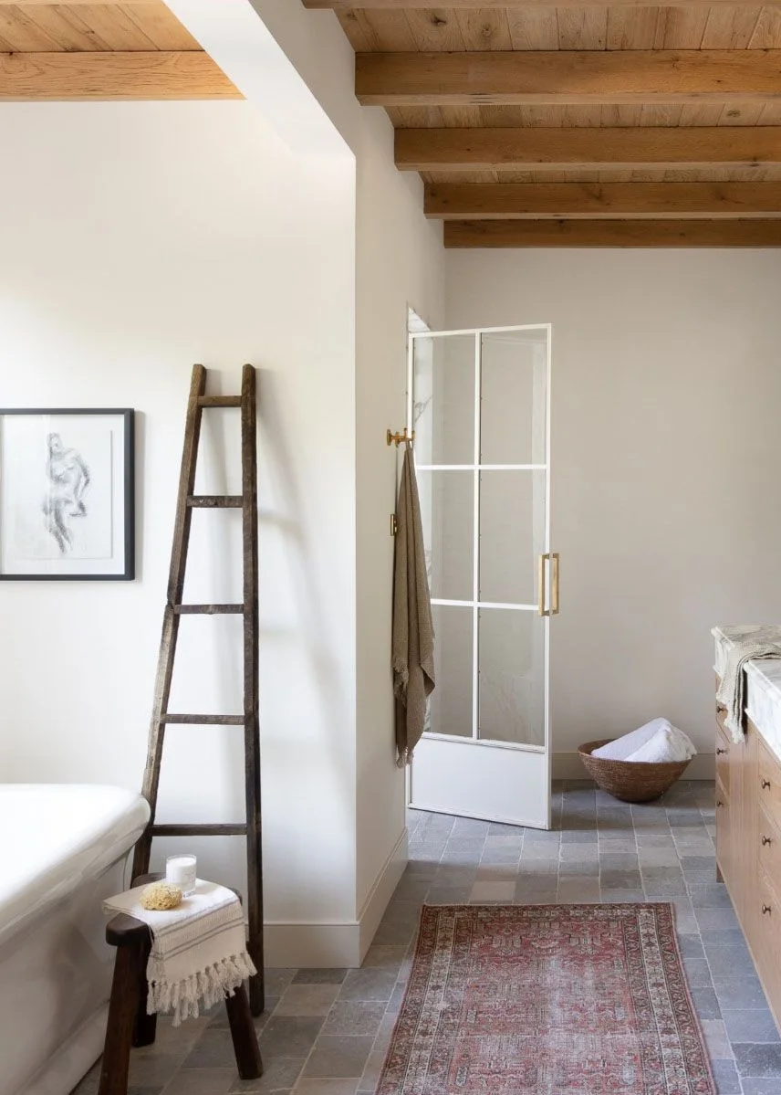 A pale bathroom with timber roof beams, a towel ladder, small stool and patterned runner
