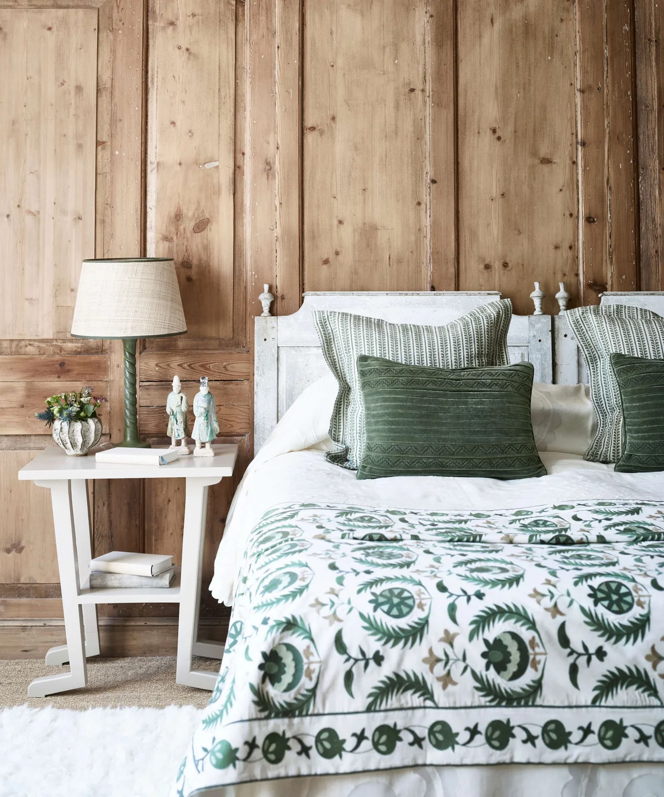 A bedroom has a broad wood-paneled wall, pale painted headboard, green patterned bedding and a small white bedside table with a green lamp.
