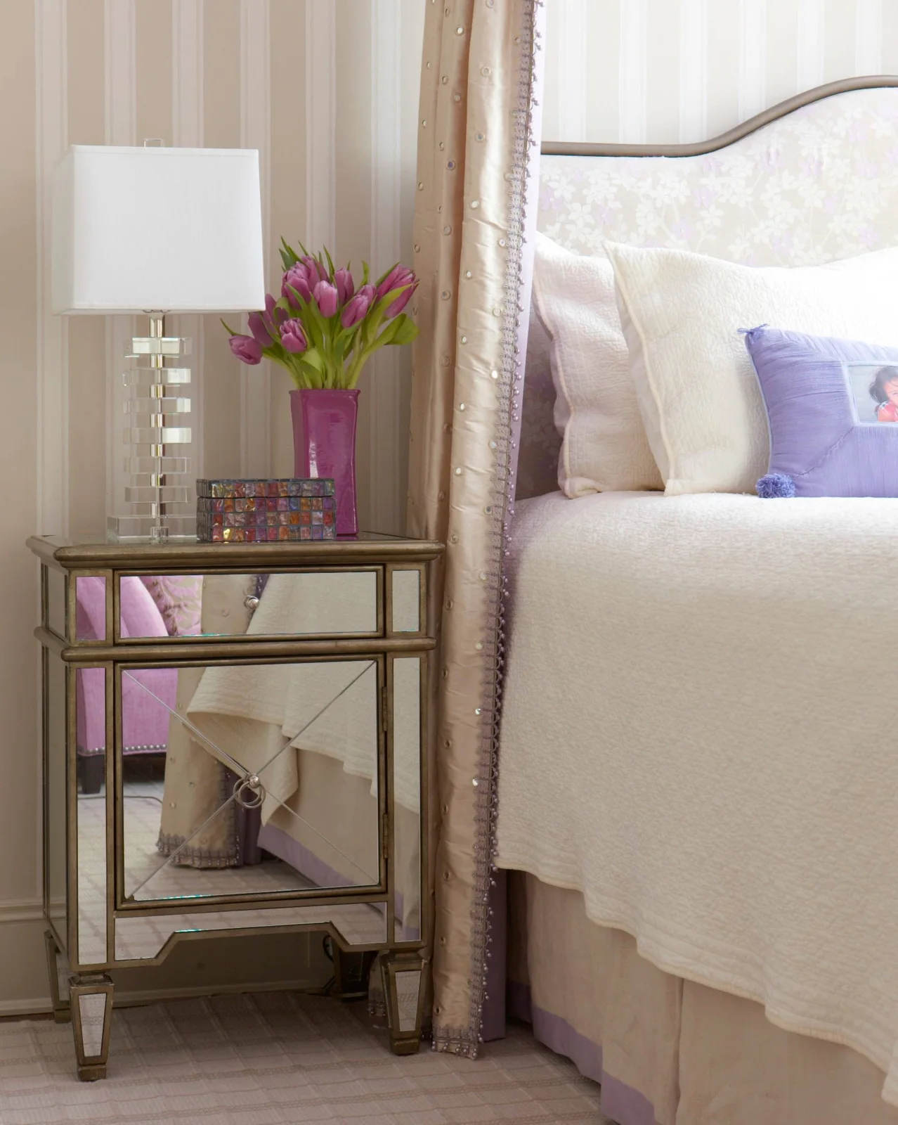 Mirrored bedside cabinet with a clear stacked lamp, purple tulips and pale upholstered bed beside a curtain