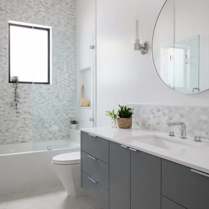 A dark gray vanity and round mirror sit beside a glass bath screen and pale small-format patterned wall tiles.