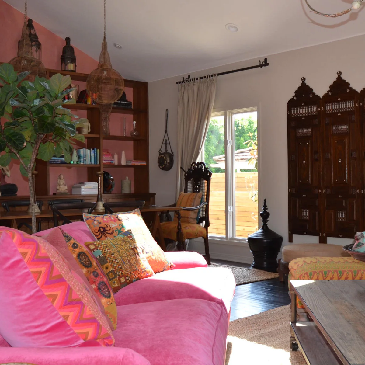 Pink sofa with patterned orange cushions beside a warm gray window wall and dark wood furnishings.