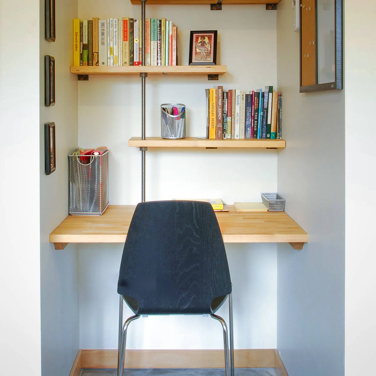 Small wood wall-supported desk beneath staggered bookshelves with a dark chair and mesh stationery holders