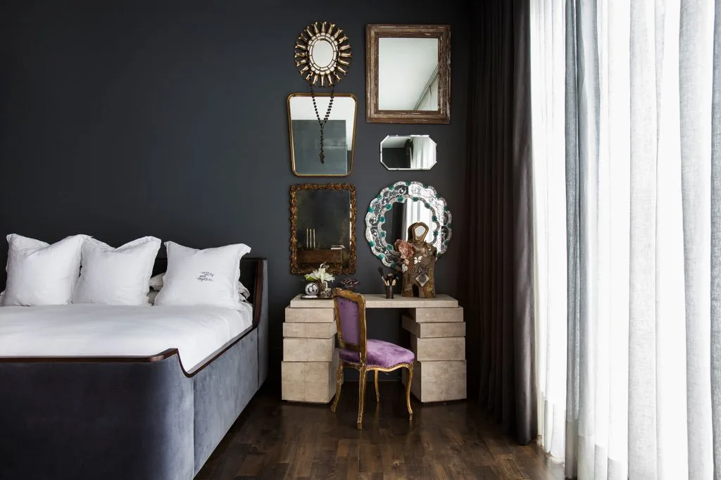 Dark bedroom wall with a mixed mirror collection above a dressing table, purple chair, pale bedding, and long light curtains.