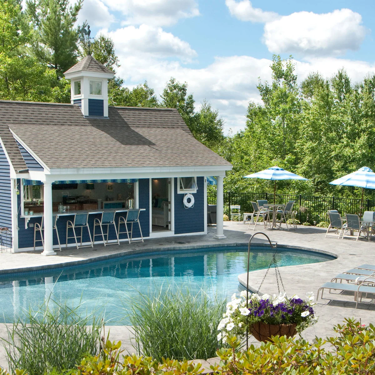 Blue pool house with a broad serving opening, four stools, white posts and separate umbrella tables beside a pool