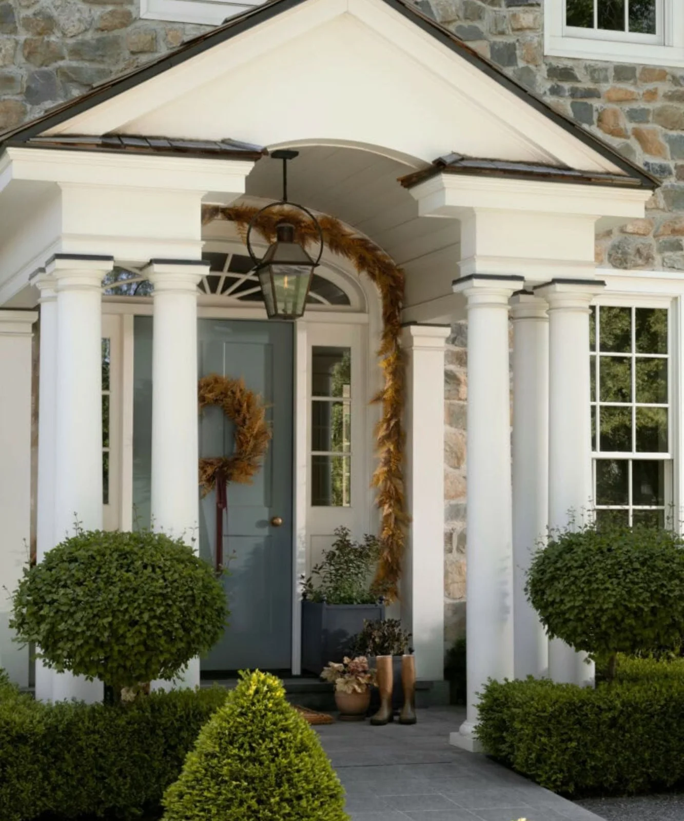 Golden-brown foliage garland frames a pale porch arch above a blue-gray door with a matching wreath and green planters